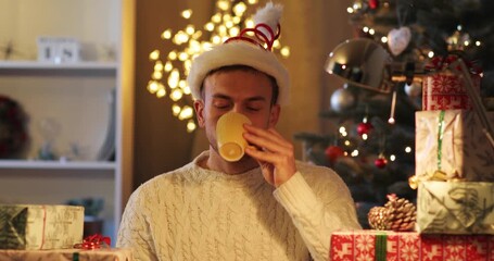 Young man with beard wearing white sweater and red funny Santa had drinking coffee. Guy sitting near gift-boxes on background of Christmas tree with decorations.