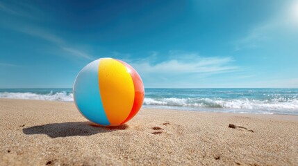 Obraz premium A vibrant beach ball lies on the warm sand near the shoreline. Waves gently crash in the background under a clear blue sky, creating a cheerful summer atmosphere.
