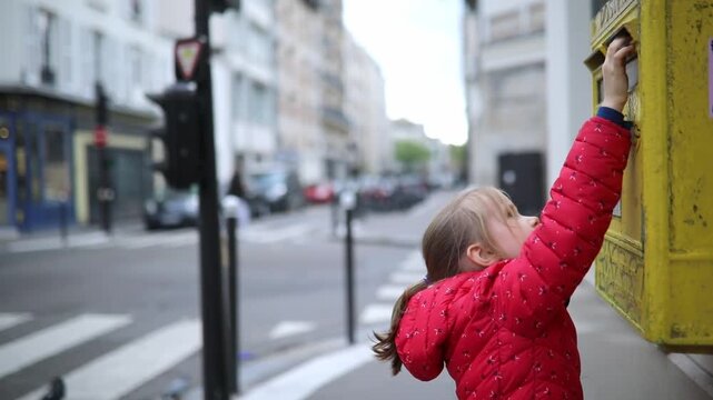 Adorable preschooler girl putting letter in yellow post box on a street of Paris