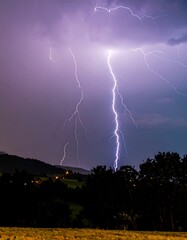 Dramatic lightning strikes over a landscape at night.  A vibrant purple-white lightning display dominates the sky, illuminating the dark hills and trees below