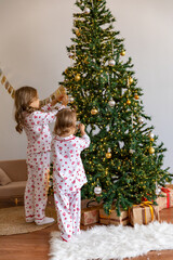 Rear view full-length little girls sisters in white pajamas with hearts print decorate Christmas tree on Christmas morning in living room at home. Happy New year family winter holiday. Copy space