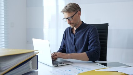 Professional businessman wearing glasses, intensely working on laptop, typing amid modern office...