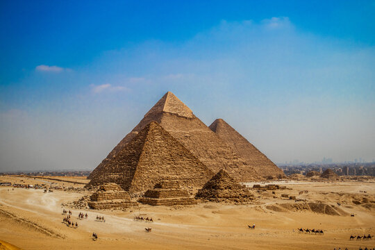 The three Great Pyramids of Giza rise above the golden desert, with long camel caravans and visitors crossing the sandy plateau while the skyline of Cairo is faintly visible on the horizon.