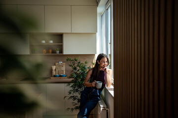Smiling woman drinking coffee and looking out the window in modern kitchen