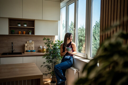 Young woman enjoying breakfast while sitting by window in modern kitchen