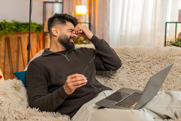 Indian man at home rubs eyes with tired expression while using laptop, fatigued from long screen exposure. Arabian guy on sofa shows discomfort and eye strain, struggling with digital overload stress