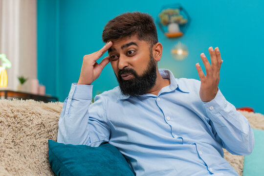 Indian man at home holds forehead facepalm after smartphone error looks disappointed regretful frustrated. Arabian guy on sofa lowers head grimaces shows guilt mistake reaction stress negative mood.