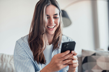 Smiling woman looking at smartphone screen, sitting on couch, enjoying leisure time with mobile device at home, happy young female chatting in social network or surfing internet, shopping online