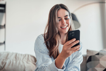Smiling woman looking at smartphone screen, sitting on couch, enjoying leisure time with mobile...