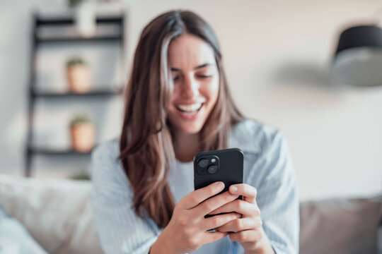 Smiling woman looking at smartphone screen, sitting on couch, enjoying leisure time with mobile device at home, happy young female chatting in social network or surfing internet, shopping online - Powered by Adobe