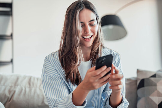 Smiling woman looking at smartphone screen, sitting on couch, enjoying leisure time with mobile device at home, happy young female chatting in social network or surfing internet, shopping online