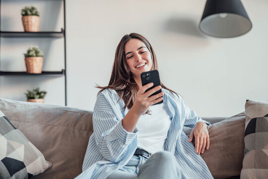 Smiling woman looking at smartphone screen, sitting on couch, enjoying leisure time with mobile device at home, happy young female chatting in social network or surfing internet, shopping online - Powered by Adobe