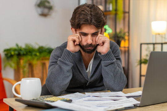 Indian man at home bends forward holds head sees unpaid bills by calculator and laptop on table. Arabian guy at desk counts fees and costs plans step low budget feels stress worry breathes slow calm.