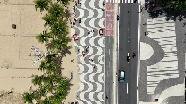 Top-down drone shot of the Copacabana mosaic pathway in Rio de Janeiro, featuring its iconic black and white stone wave design, rows of palm trees, wide sandy shoreline, and the vibrant beachfront atm