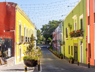 A street with two buildings, one yellow and one green. The street is lined with potted plants and has a few cars parked along it. City of Puebla, Mexico, crafts
