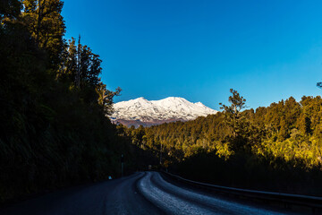 A scenic view of Mount Ruapehu covered in snow, framed by dense forest trees and a winding road in...