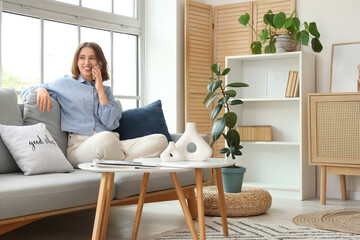 Pretty young woman with mobile phone sitting on grey sofa near window in light living room