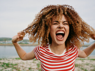 Close-up of a joyful young woman shouting with arms raised, curly hair bouncing, wearing a red and...