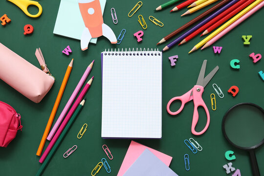 Blank notebook with different stationery supplies and paper rocket on green chalkboard