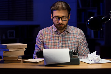 Young male writer with books and vintage typewriter working at home in evening