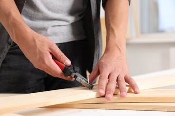 Man pulling metal nail out of wooden plank with pincers indoors, closeup