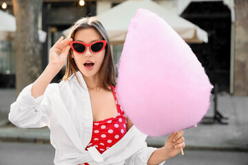 Beautiful young shocked woman in sunglasses with cotton candy on city street, outdoors
