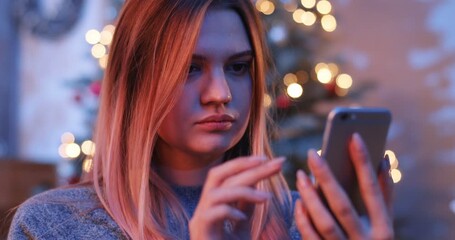 Confused pretty girl dealing the settings of mobile phone. Young woman holding smartphone in hands. Close-up. Winter. Evening.
