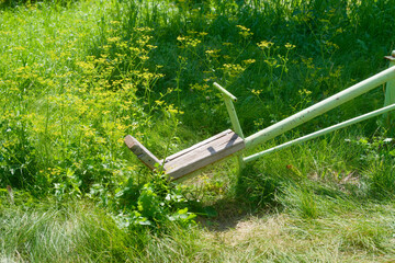 Fragment of an old retro swing seat with wooden board among tall green grass in summer garden,...