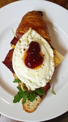 Delicious Croissant Sandwich with Fried Egg, Bacon, Cheese, and Arugula on a White Plate