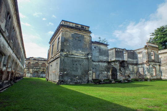 A panoramic view of the Van den Bosch Fortress in Ngawi, East Java, showing its weathered Dutch colonial architecture and ruined buildings surrounded by a vast green lawn under a cloudy sky.