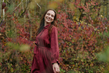 A candid portrait of a smiling woman in a wine red dress amid autumn foliage, conveying authenticity and credible presence through natural lighting and relaxed pose.