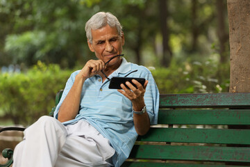 Indian old man looking mobile phone at the park. Senior man watching video in phone. Aging man reading online news through mobile phone.