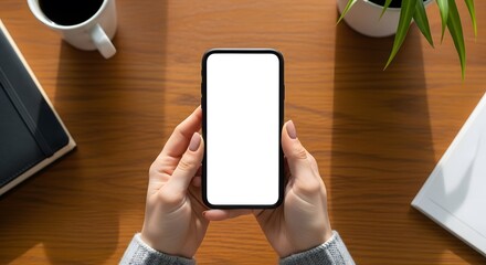 Overhead view of hands holding a contemporary smartphone with a blank white screen, perfectly framed on a wooden desk with office essentials, ready for customization