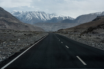 An empty asphalt road leading through barren rocky terrain toward snow-covered mountains under a cloudy sky symbolises travel, adventure, and remote landscapes.