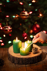 A hand ignites a green candle with a burning match. Candles sit on a wooden surface near pine cones with a decorated Christmas tree and garland in the background.