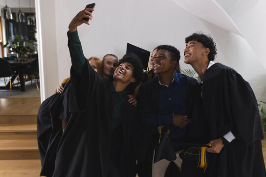 Graduating teens in caps and gowns taking selfie, celebrating together indoors