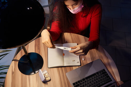 sick woman with pills and thermometer on a table working on a laptop from home on quarantine isolation.
