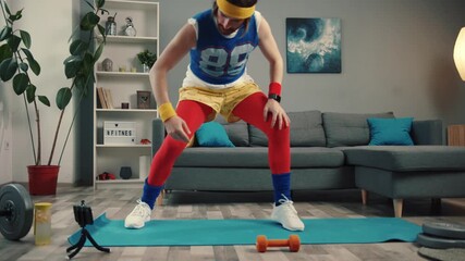 LOS ANGELES, CALIFORNIA, 10 OCTOBER, 2019: Funny-looking retro fitness instructor shooting exercise videos for vlog at home. Concentrated young man stretching legs and warming up before training.
