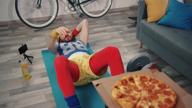 Weak funny stylish sportsman trying to do abs crunches on mat and taste delicious hot pizza. Ab training and home workout.