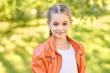 Caucasian female child smiling in orange jacket outdoors with green leafy background on a sunny day.