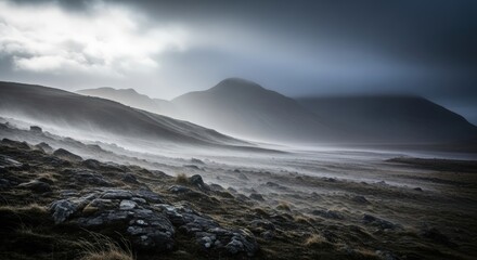 Fototapeta premium Misty mountains shrouded in atmospheric fog and low clouds, with rugged terrain in the foreground.