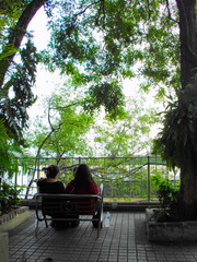 Two people sit closely on a bench beneath leafy trees, gazing at the view ahead, captured in soft retro tones with film grain.
