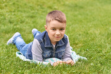 Young caucasian child in denim vest lying on green grass smiling outdoors on a sunny day.