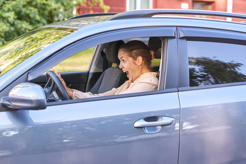 Surprised caucasian adult female driver in blue car reacting to unexpected situation on sunny day.