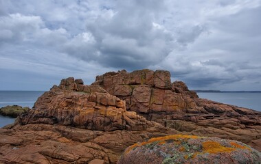 Massive reddish coastal cliffs covered with moss and lichen under a dramatic cloudy sky. Wild seashore landscape with textured rocks and ocean in the background. France