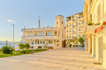 A sunny promenade in a coastal town, with modern buildings and a view of the sea and mountains. The golden hour light creates a serene and inviting atmosphere.