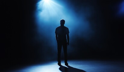 Man stands on stage under spotlight facing empty dark room with no people around him illuminated by blue light from above casting long shadows down onto ground below him