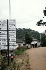 This image shows a rural road junction with a prominent signpost listing various local destinations in both Thai and English.