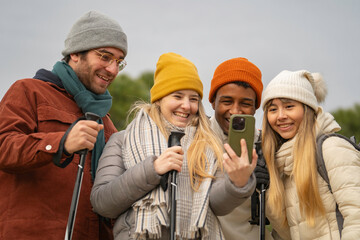 Multiethnic group of four cheerful hikers taking a selfie with a smartphone while enjoying a nordic walking excursion in winter