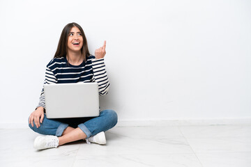 Young caucasian woman with a laptop sitting on the floor isolated on white background pointing up and surprised